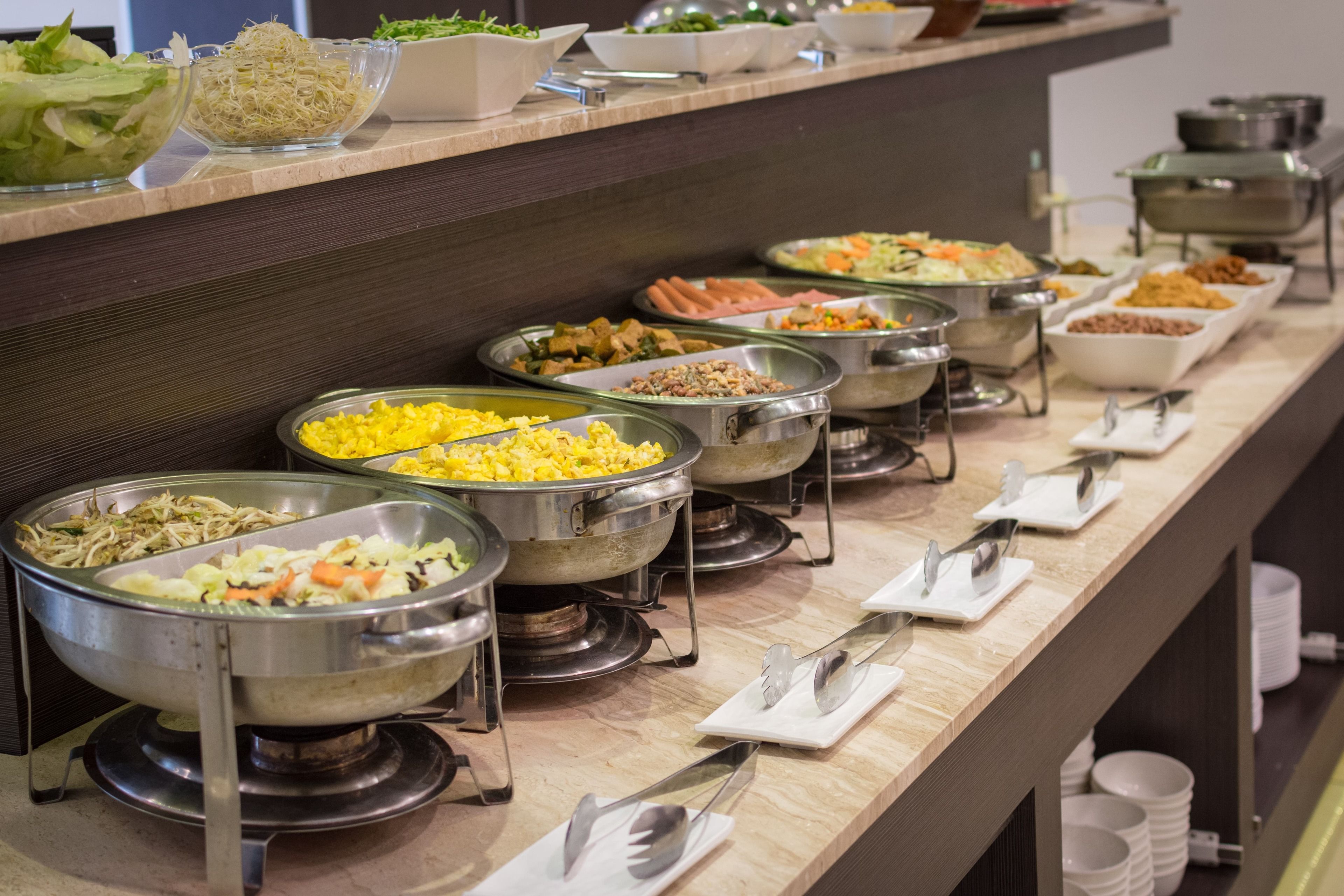 Een buffet met verschillende soorten eten in metalen schalen en bakplaten, bestek op witte servetten en een houten toonbank.