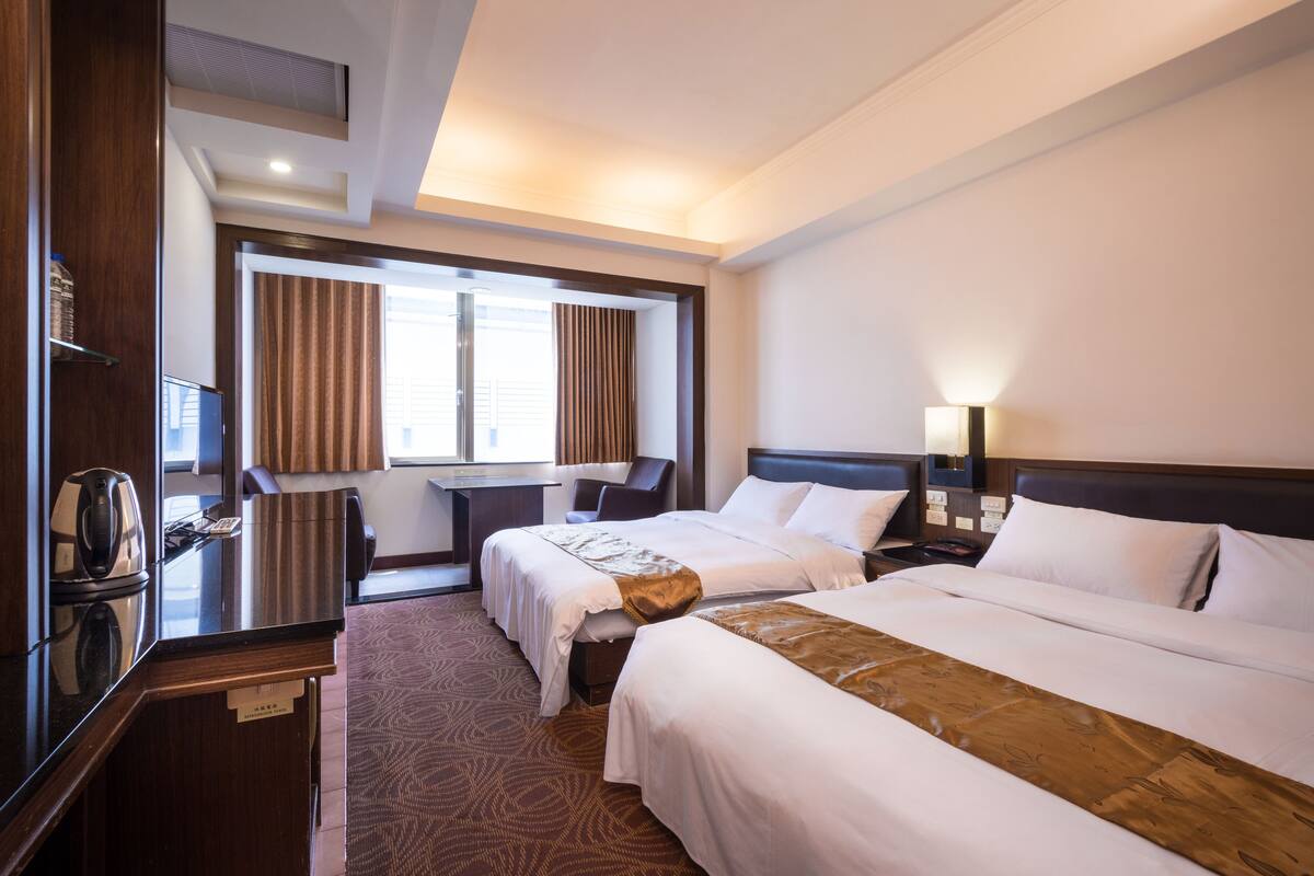 A hotel room with two beds, a desk, a chair, and a window with curtains.