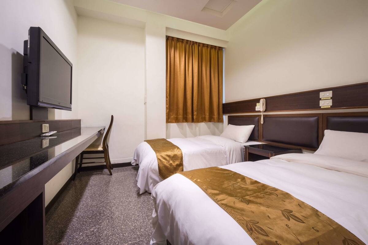A hotel room with two beds, a desk, a television, and a window with curtains.