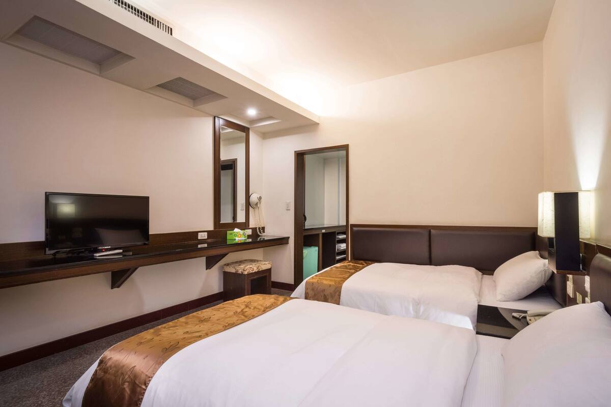 A hotel room with two beds, a TV, a desk with a mirror, and a bedside lamp.