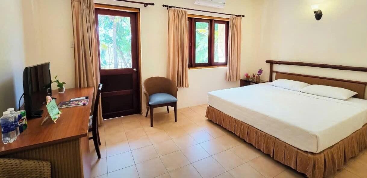 Une chambre d’hôtel avec un lit, un bureau avec un ordinateur, une chaise et des fenêtres avec des rideaux.