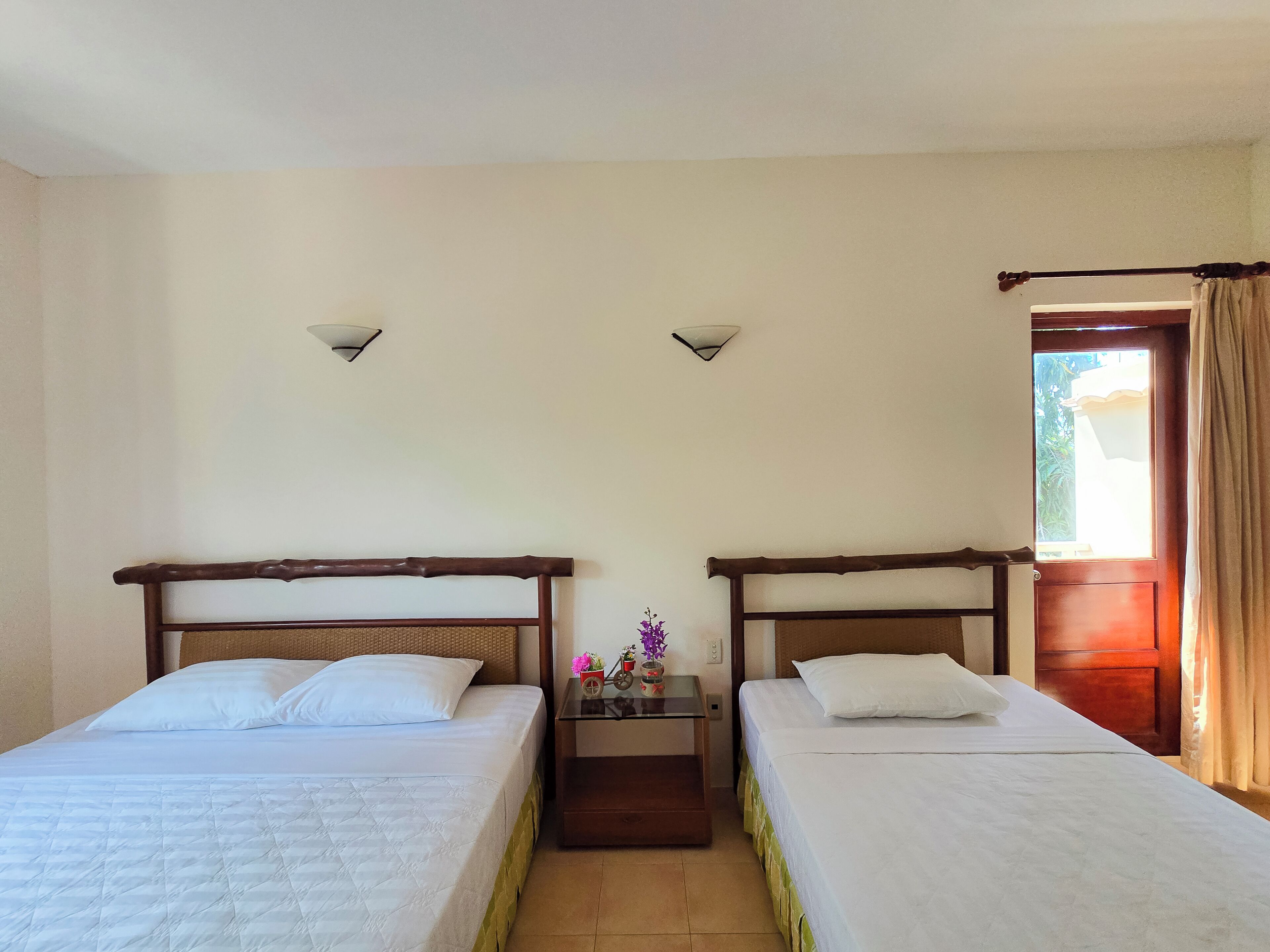 Ein Zimmer mit zwei Betten, einem Nachttisch mit einer Vase und einer Tür mit Vorhängen.