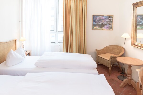 Une chambre d’hôtel avec deux lits, une chaise en osier, une petite table ronde et un tableau au mur.