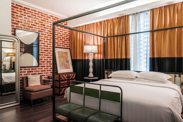 Una cama con dosel de cuatro postes con estructura metálica verde, colcha blanca y cortinas negras y anaranjadas. Se ve una silla de mimbre y una mesita con una lámpara.