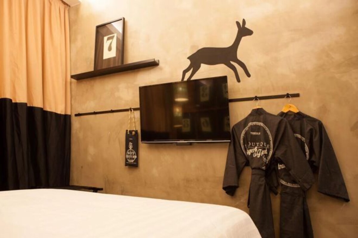 A hotel room with a bed, a mounted TV, a wall-mounted shelf with a picture frame, and a wall decoration of a deer.