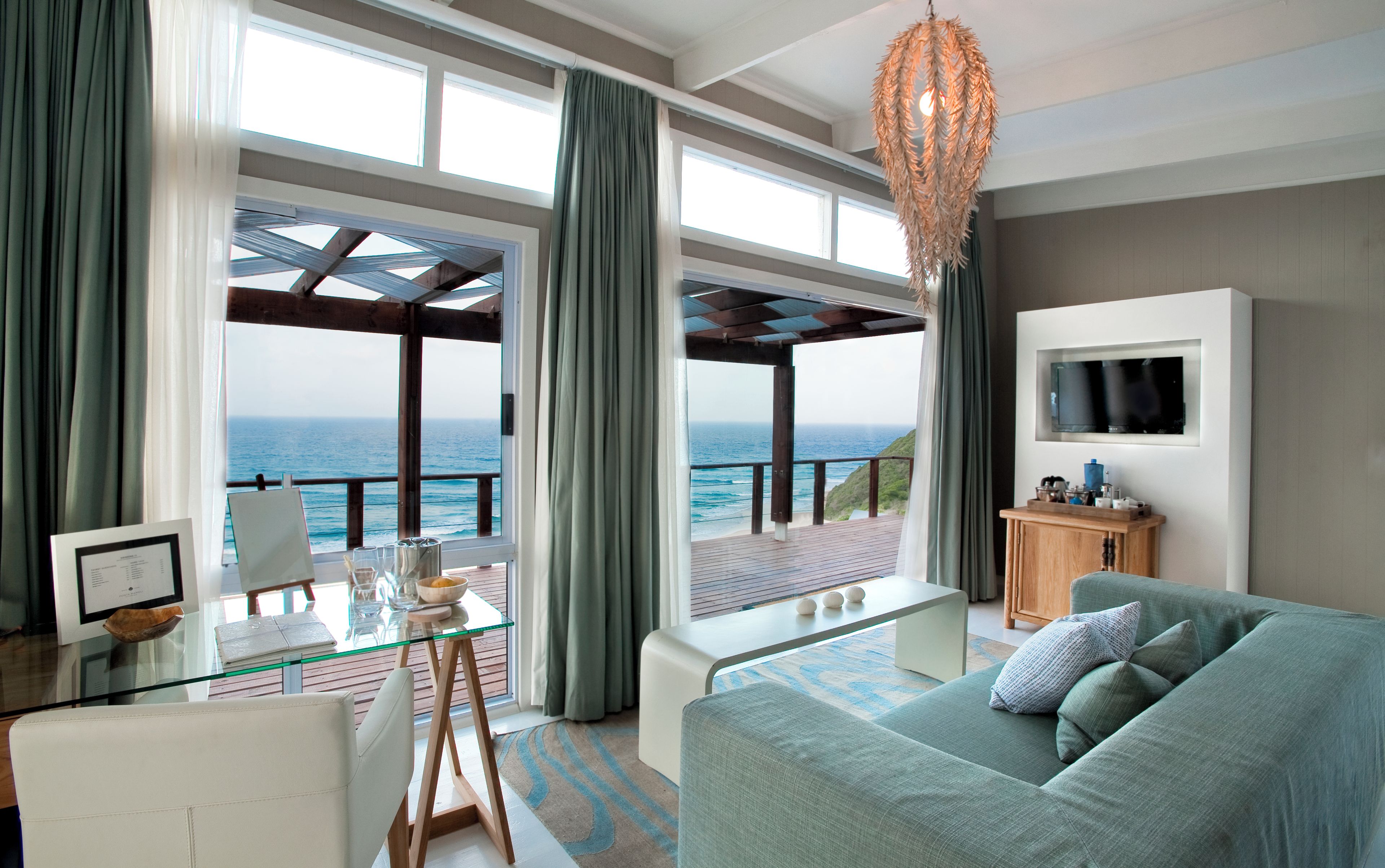 Una sala de estar moderna con un amplio ventanal con vistas al mar, una mesa de vidrio, un sofá y un televisor montado en la pared.