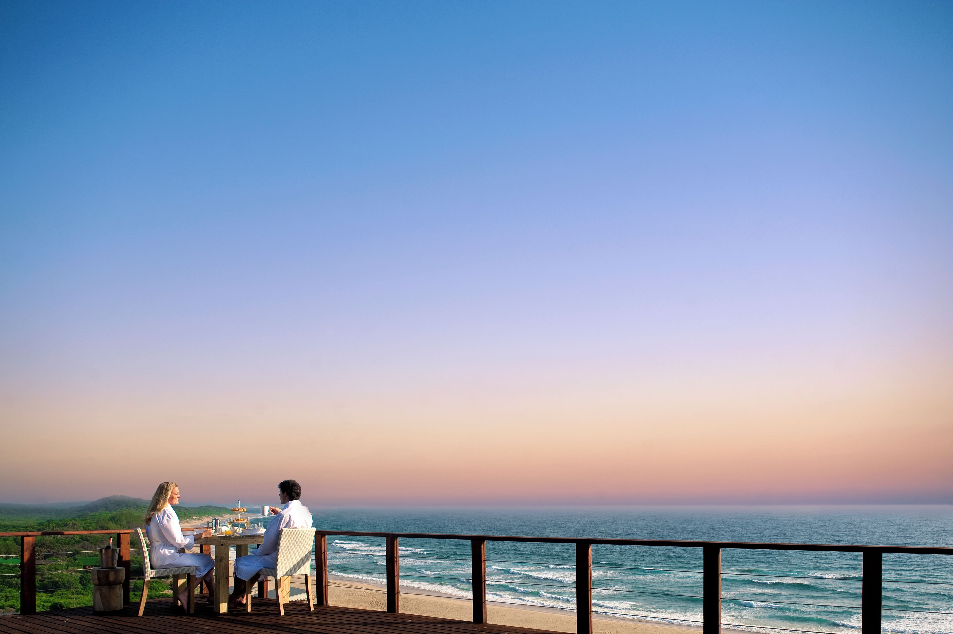 A couple dining at a wooden deck overlooking a beach with a view of the ocean and distant hills.