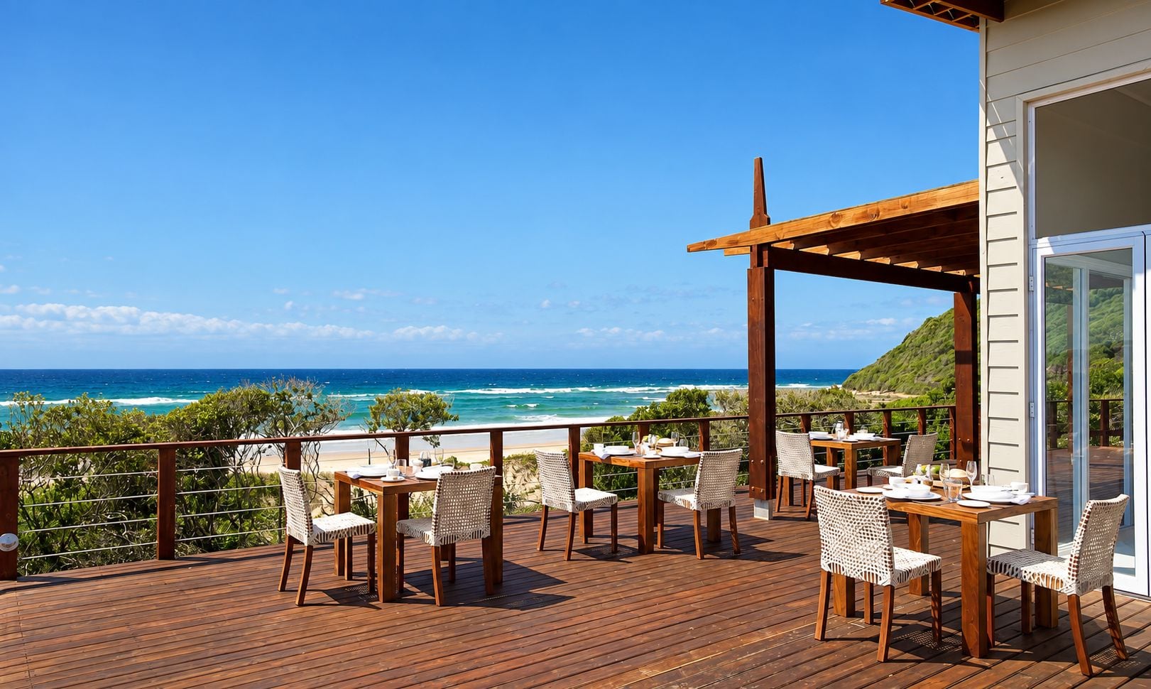 Eine Holzterrasse mit Tischen und Stühlen, die auf einen Strand und das Meer blickt.