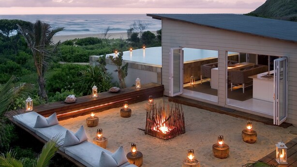 A modern outdoor lounge area with a fire pit, seating, and lanterns, overlooking a beach and ocean. - White Pearl Resorts (Zitundo)