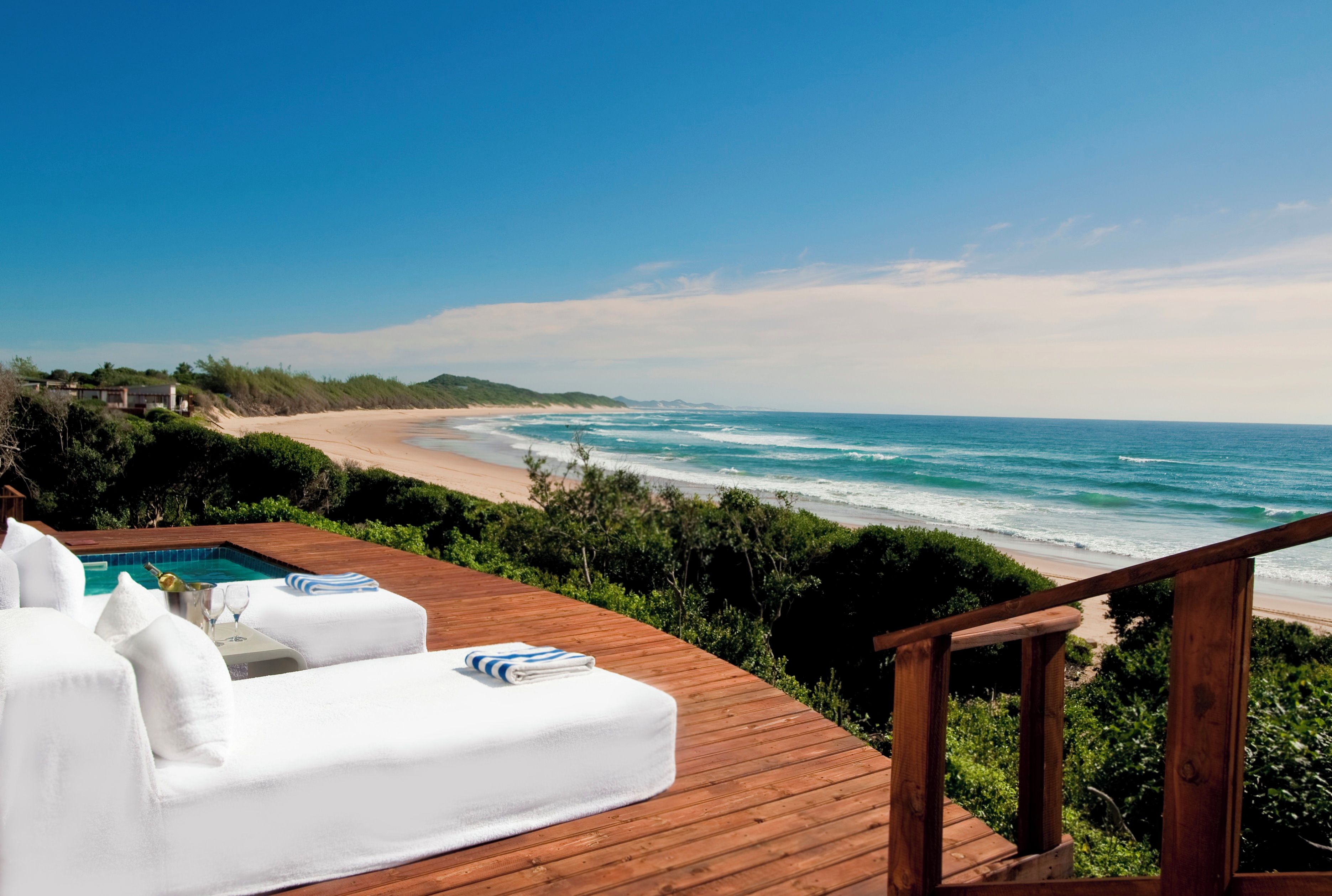 Una terraza de madera con piscina, sillas de descanso blancas y vistas a la playa y el océano.