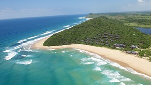 An aerial view of a coastal area with a beach, a forested hill, and several buildings. - White Pearl Resorts (Zitundo)