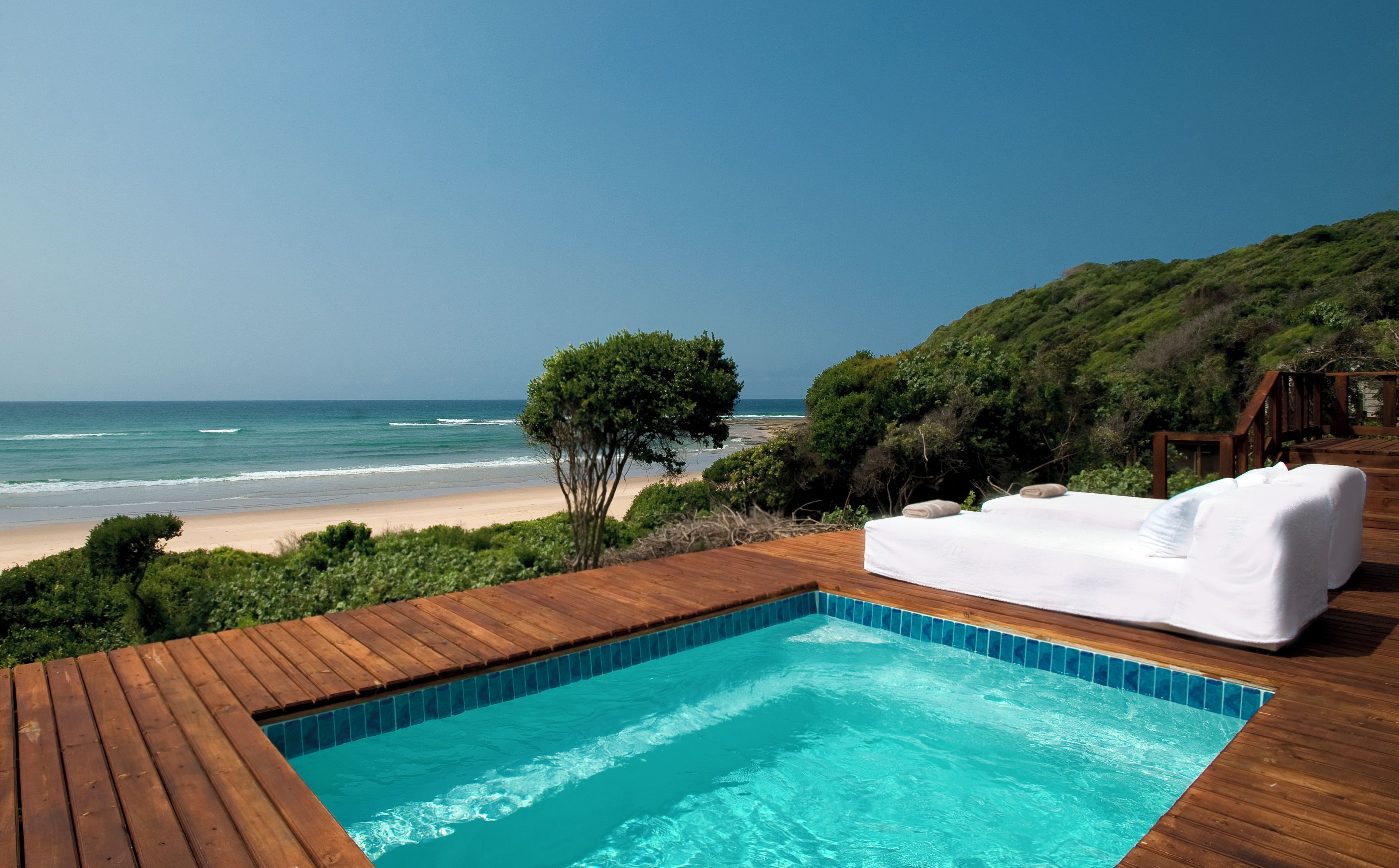 A wooden deck with a swimming pool, a white outdoor sofa, and a view of the beach and ocean.