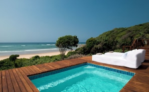 A wooden deck with a swimming pool, a white outdoor sofa, and a view of the beach and ocean.