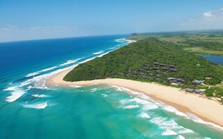 Vista aérea de una zona costera con playa, océano y una colina boscosa con edificios.