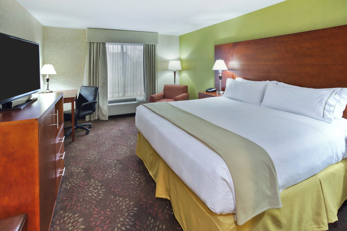 A hotel room with a large bed, a desk with a TV, a chair, and a window with curtains.