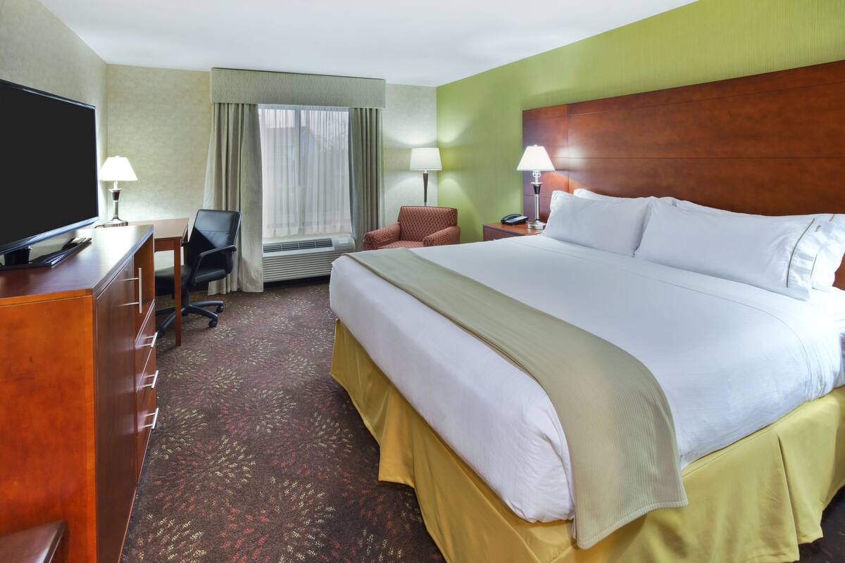A hotel room with a large bed, a desk with a TV, a chair, a lamp, and a window with curtains.