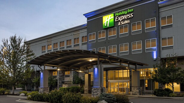 A Holiday Inn Express & Suites building with a covered entrance, stone pillars, and a landscaped area with shrubs and a bench. - Holiday Inn Express & Suites Colorado Springs Central by IHG (Colorado Springs)