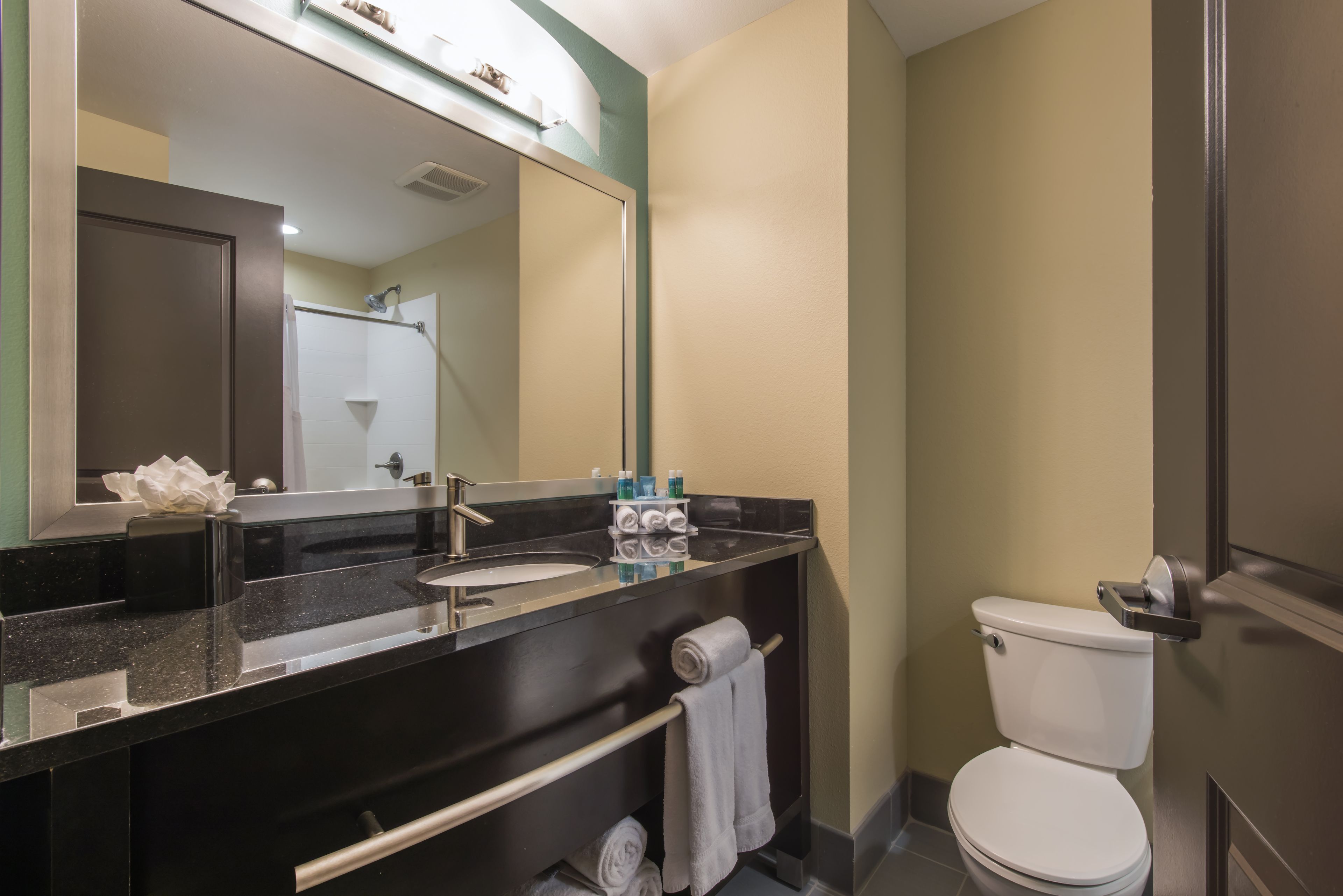 Un baño moderno con lavabo negro, espejo y inodoro blanco. Hay toallas y artículos de aseo en la encimera.