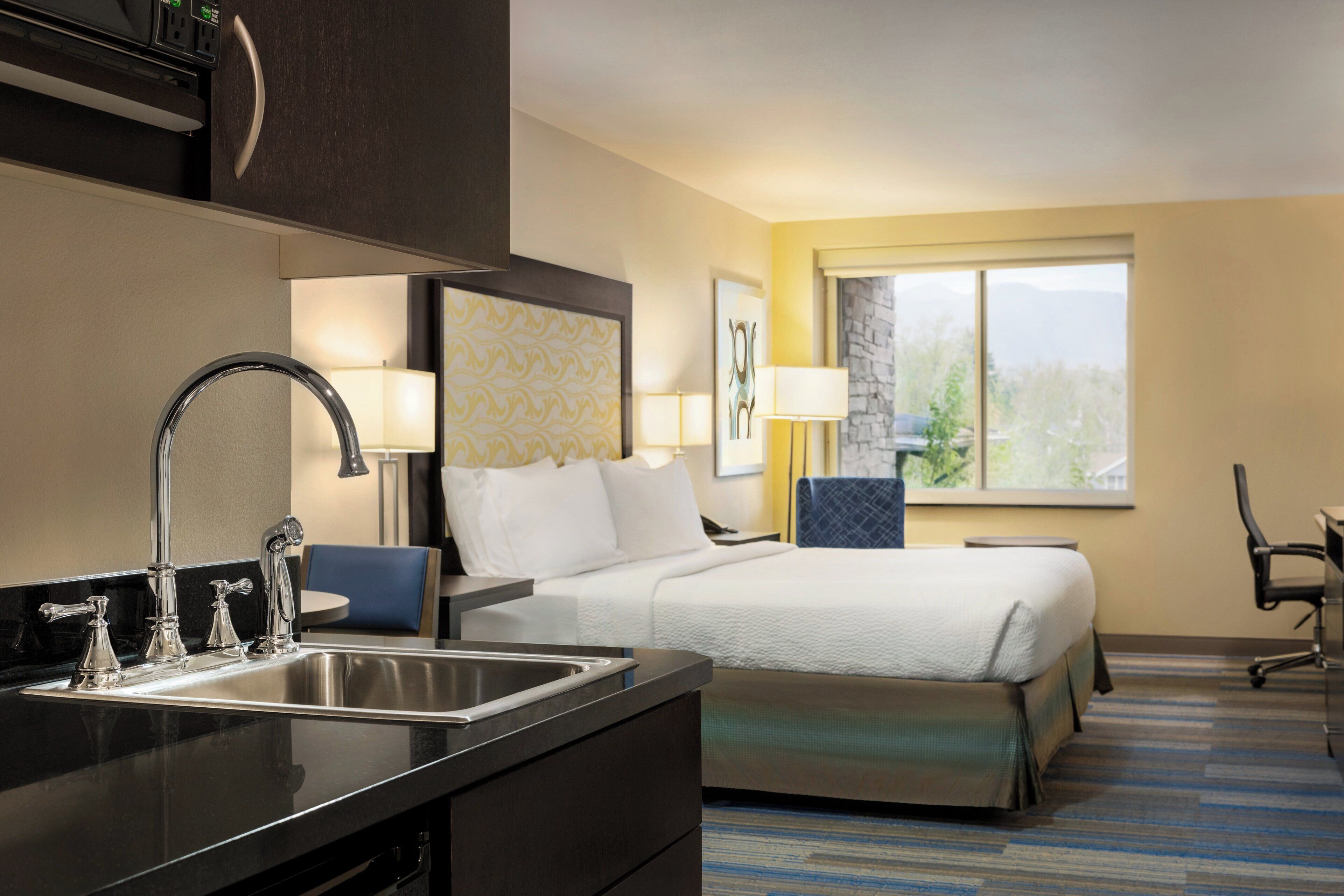 Habitación de hotel con cama, escritorio, silla, lavabo y ventana con vistas a árboles.