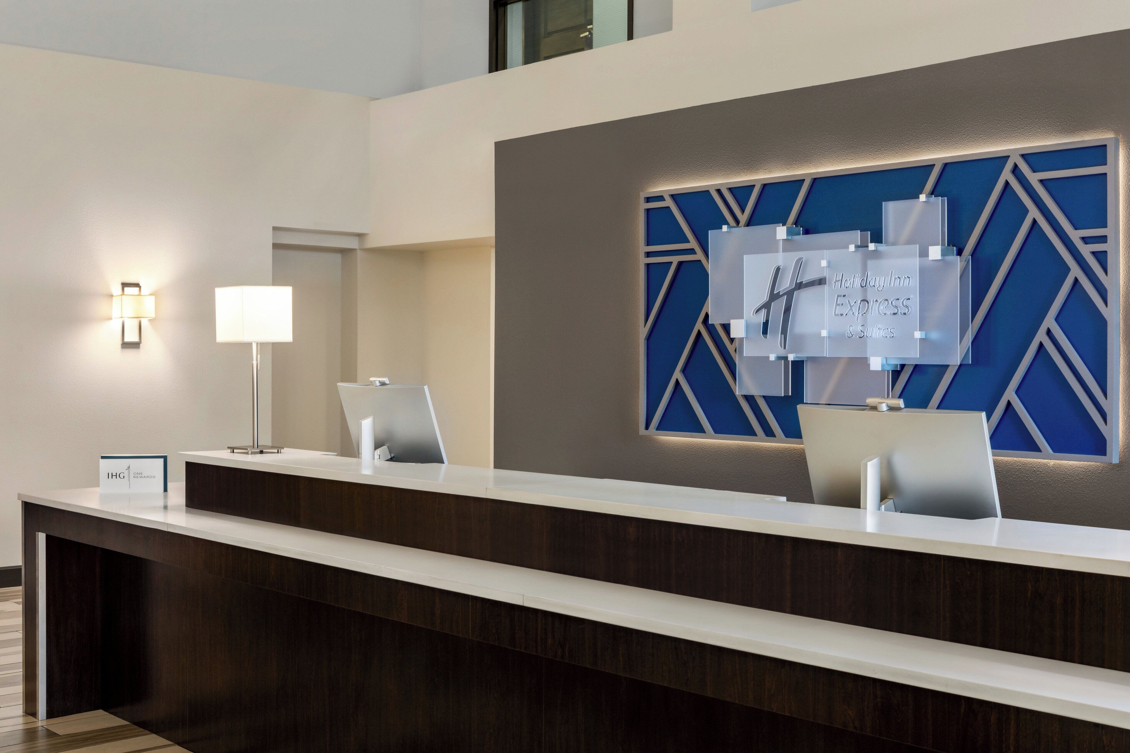 Un moderno mostrador de recepción con pantalla digital, lámpara de escritorio y un cartel con el nombre del hotel.