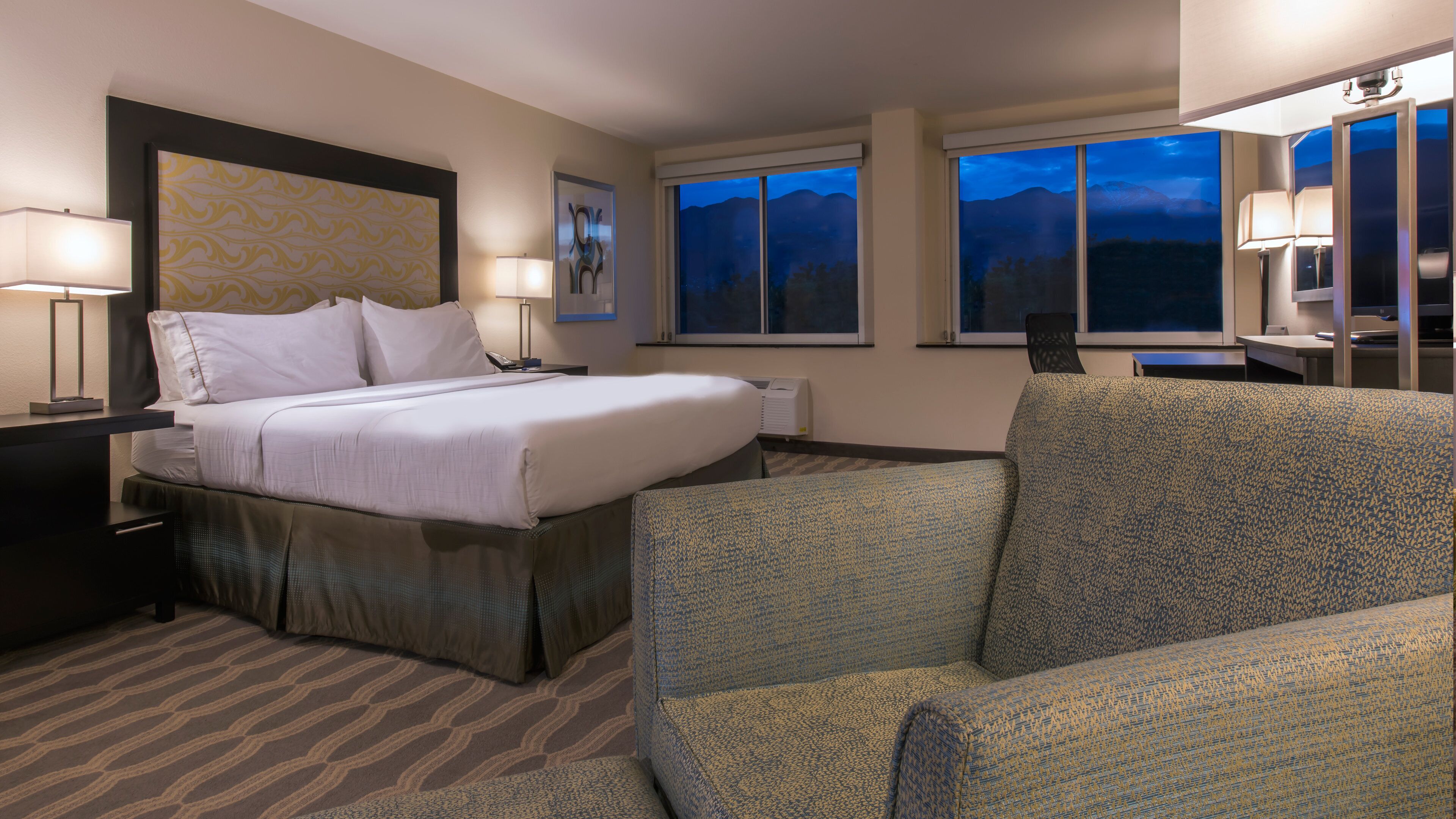 A hotel room with a bed, two bedside lamps, a desk, a chair, and a large window with a view of mountains.