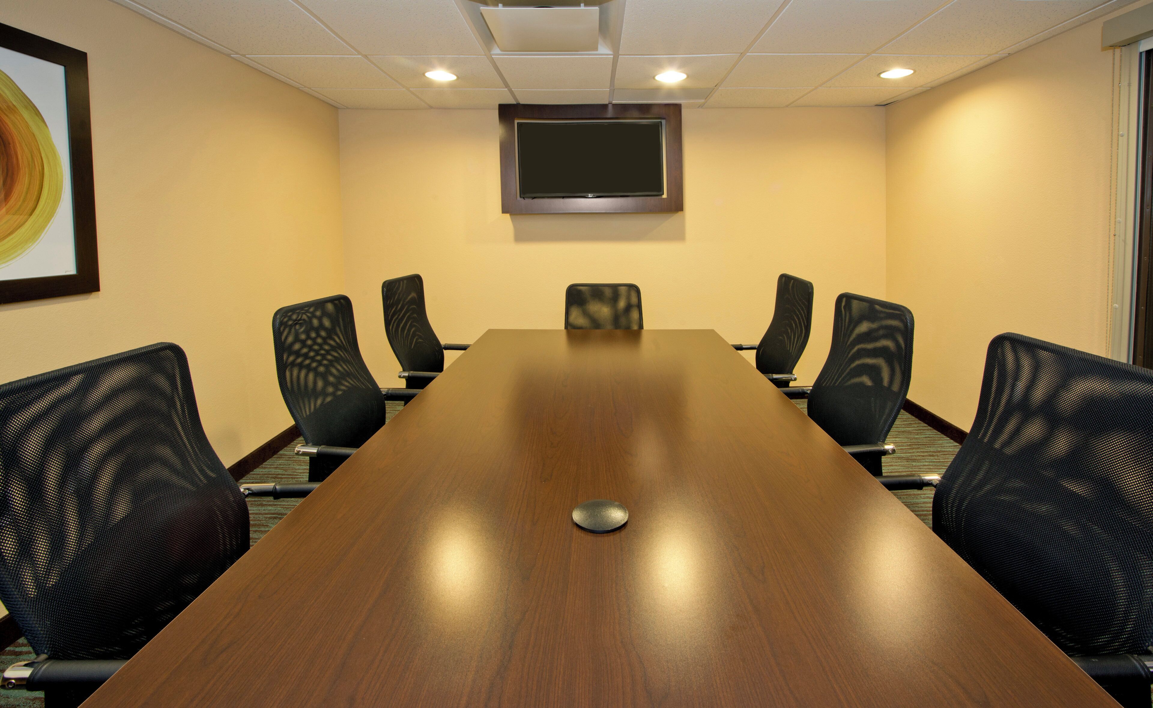 A conference room with a long wooden table, six chairs, a flat-screen TV mounted on the wall, and a framed artwork on the wall.
