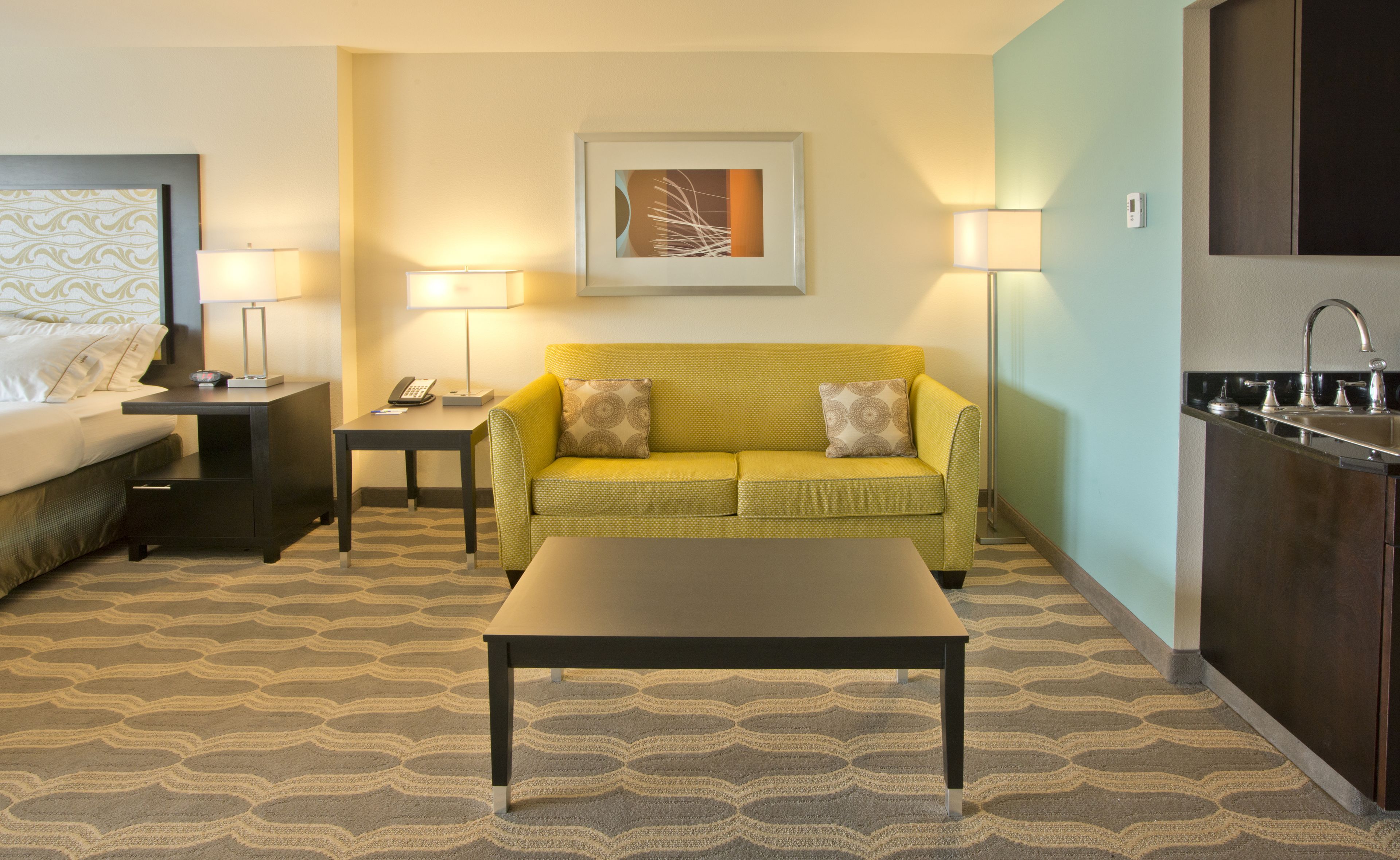 A hotel room with a bed, a yellow sofa, a coffee table, a desk with a phone, a lamp, a painting on the wall, and a kitchen area with a sink.