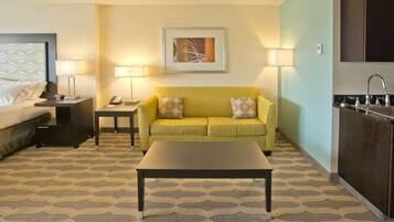 A hotel room with a bed, a yellow sofa, a coffee table, a desk with a phone, a lamp, a painting on the wall, and a kitchen area with a sink.