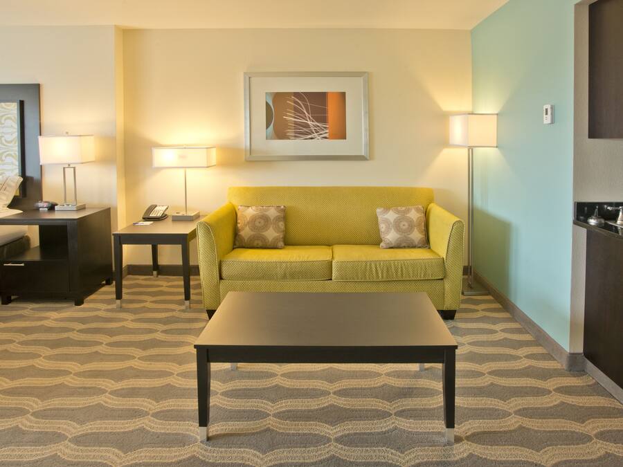A hotel room with a bed, a yellow sofa, a coffee table, a desk with a phone, a lamp, a painting on the wall, and a kitchen area with a sink.