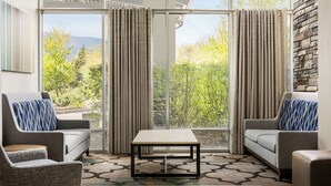 A modern living room with a sofa, armchair, and coffee table, large windows with curtains, and a stone wall. - Holiday Inn Express & Suites Colorado Springs Central by IHG (Colorado Springs)