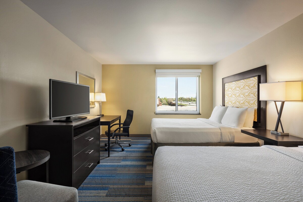 A hotel room with two beds, a desk with a TV, a chair, a window, and a lamp.