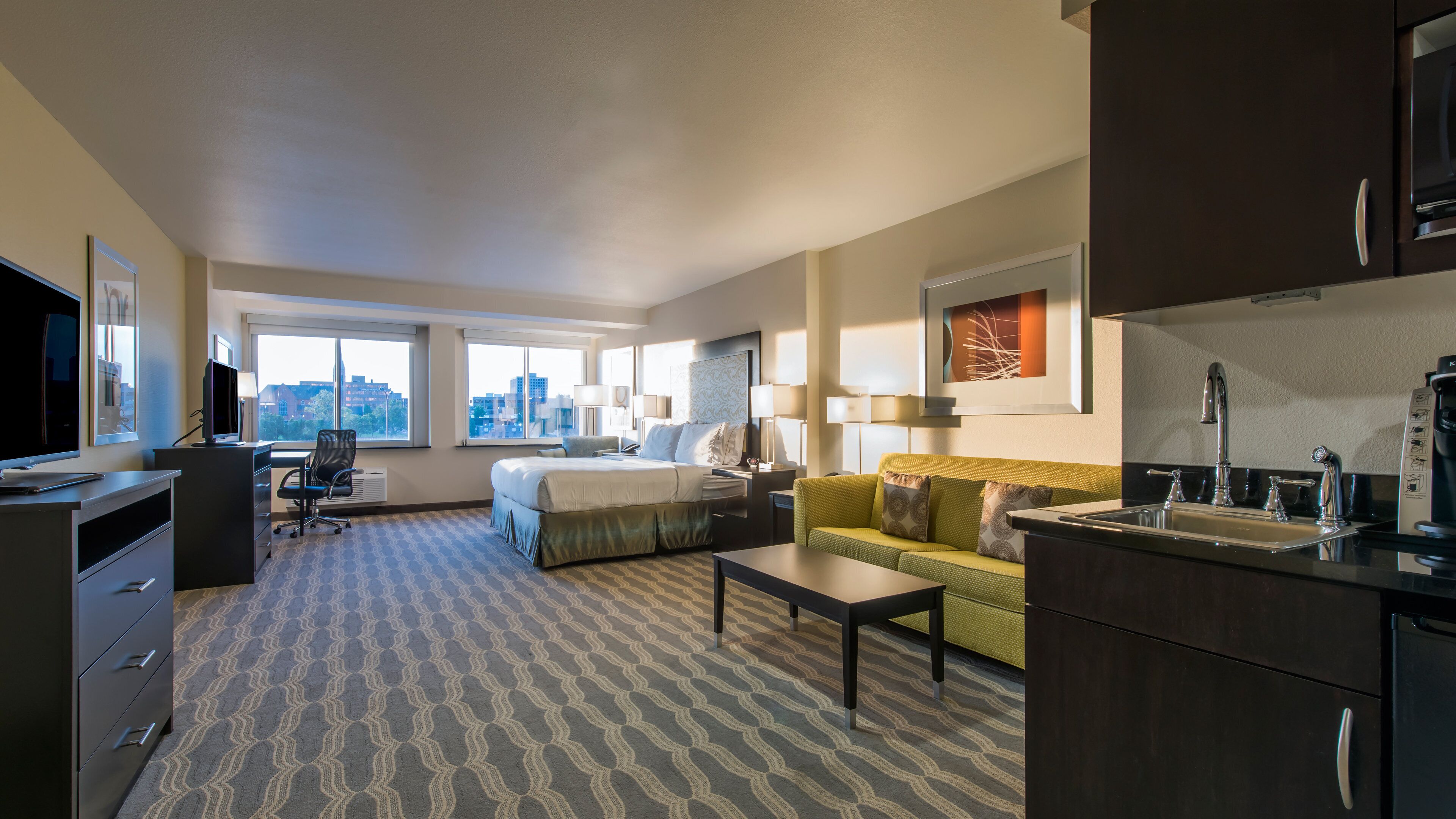 A hotel room with a bed, a sofa, a desk with a computer, a television, and a city view.