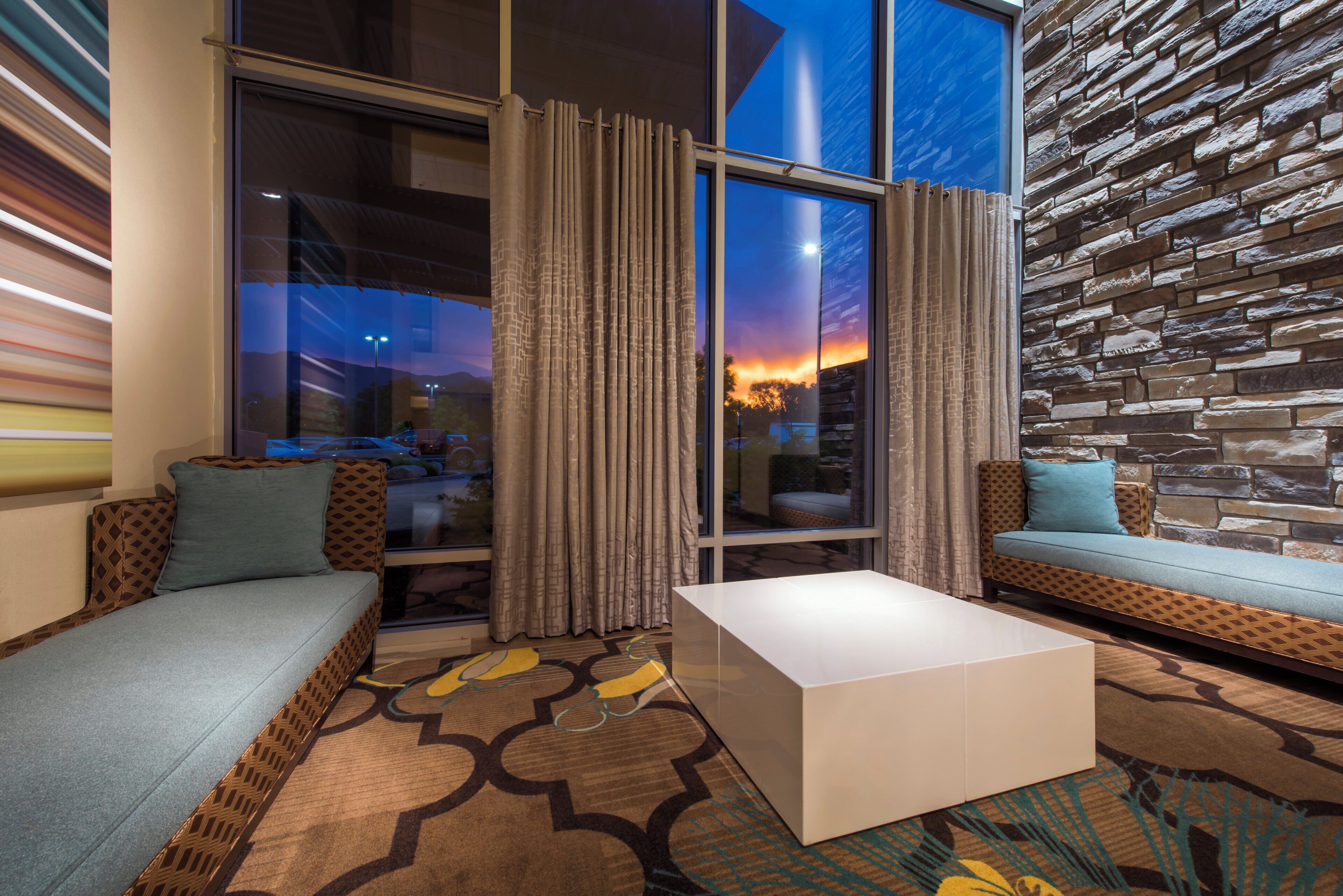A modern hotel room with a large window, a patterned rug, a white coffee table, and a stone wall.