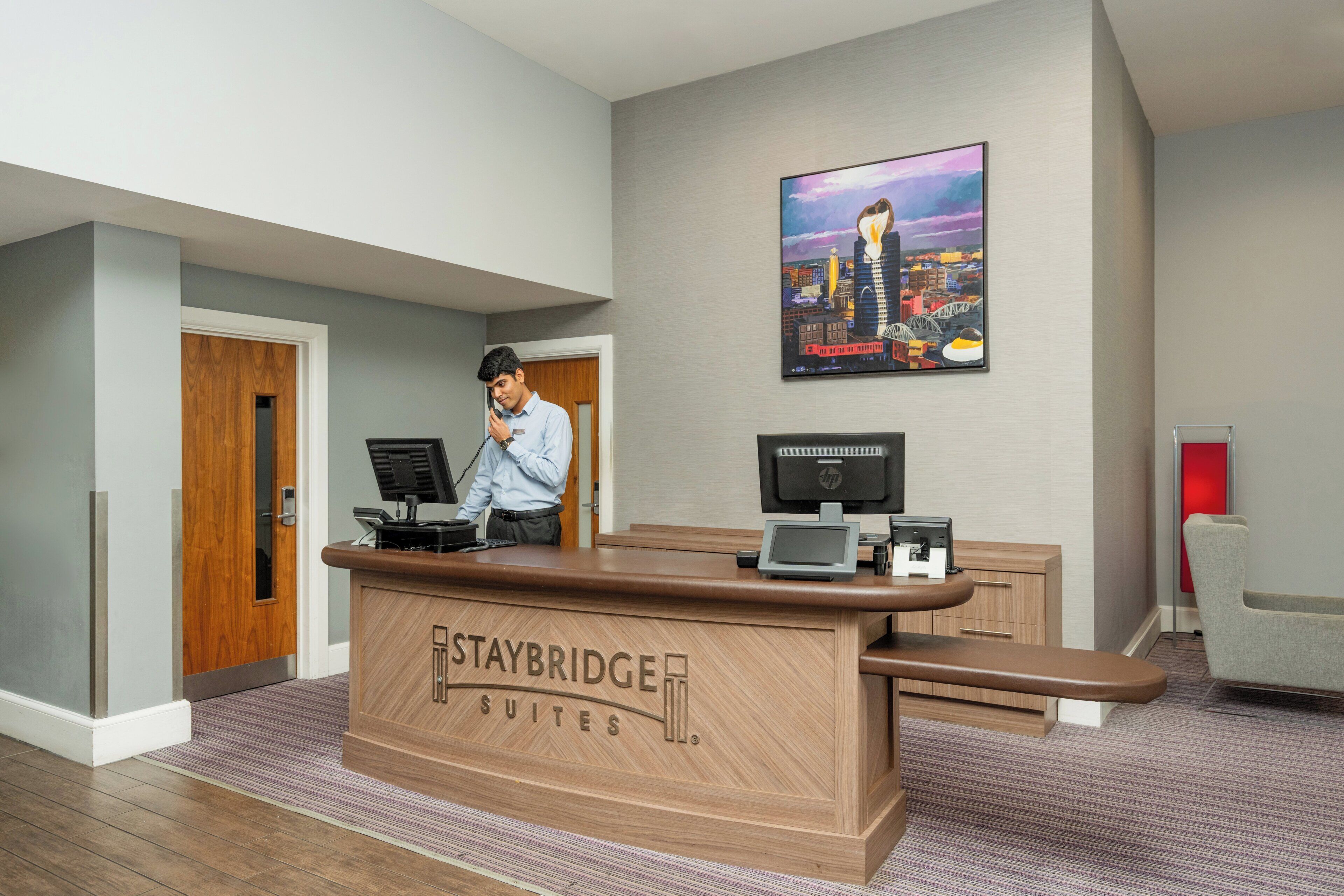 Un comptoir d’accueil avec une enseigne indiquant « Staybridge Suites » et un tableau représentant un paysage urbain accroché au mur.