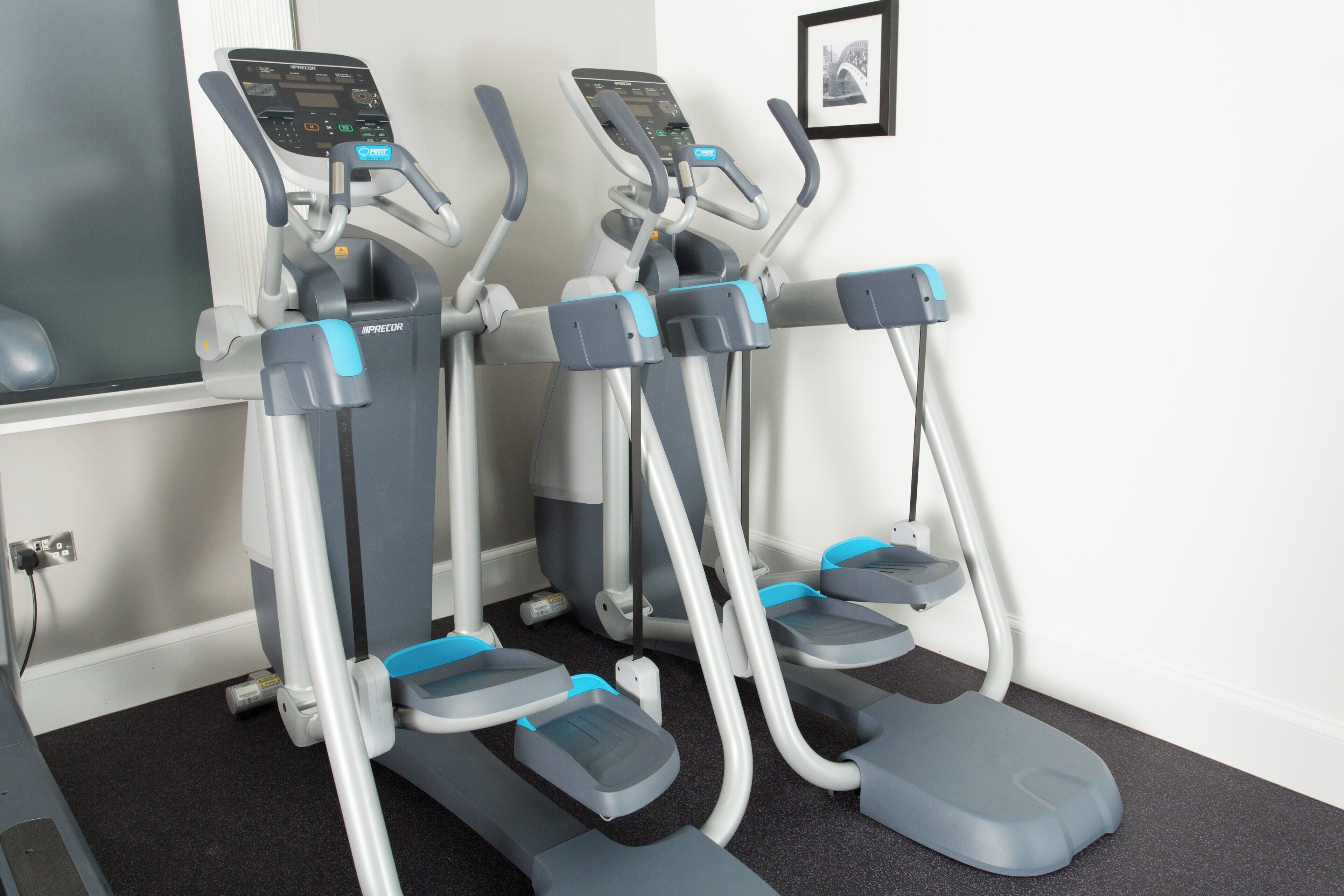 Three exercise machines with digital displays, positioned against a wall with a framed picture.