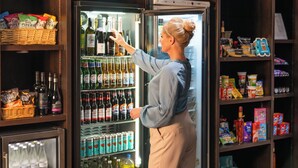 Una mujer en un pasillo de tienda frente a un expositor refrigerado con diversas bebidas y aperitivos.