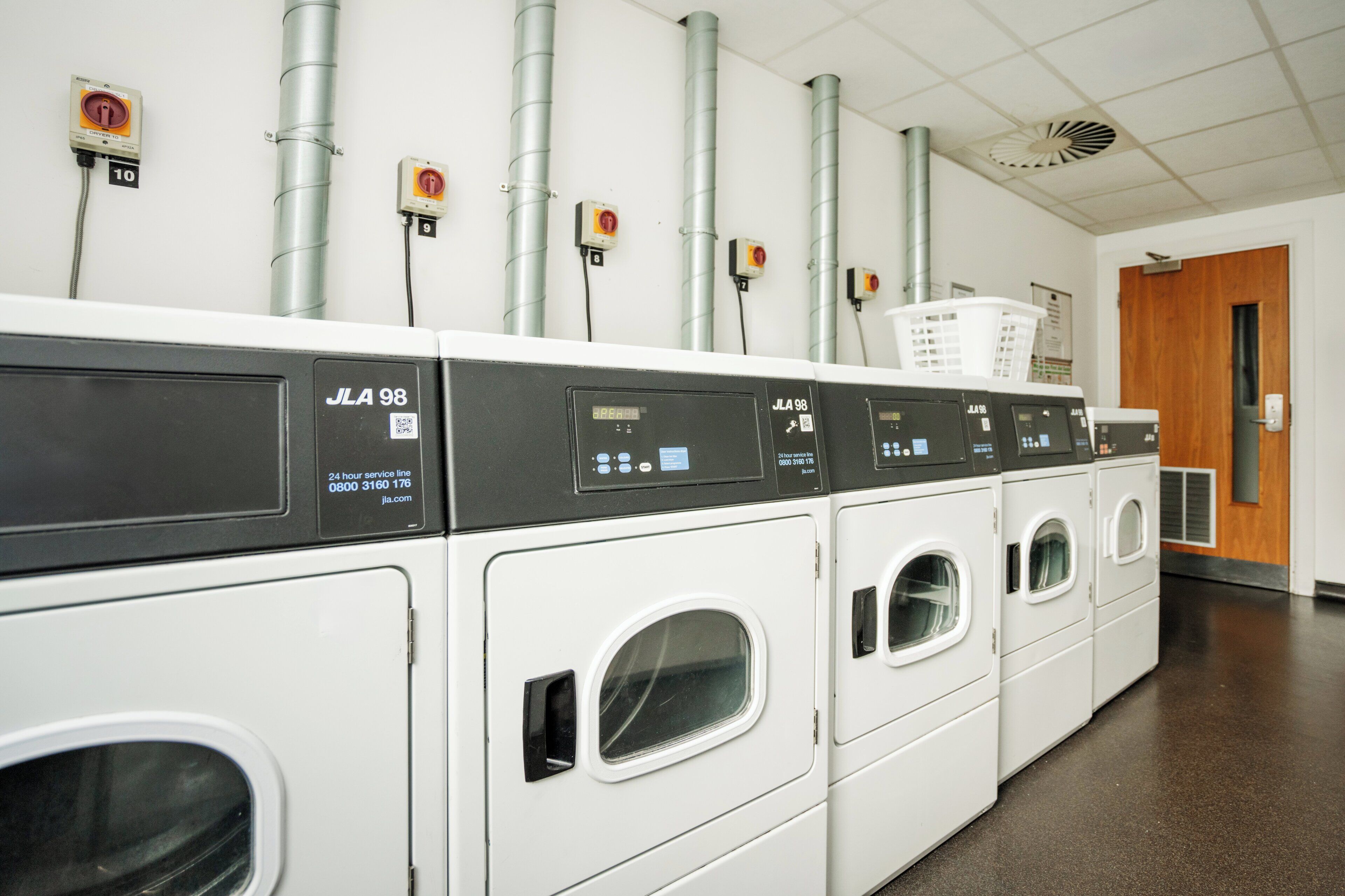 A row of white front-loading washers with control panels, connected to overhead pipes and safety switches on the wall.