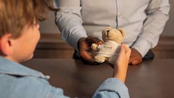 A child holding a teddy bear with a label, sitting at a table with an adult.