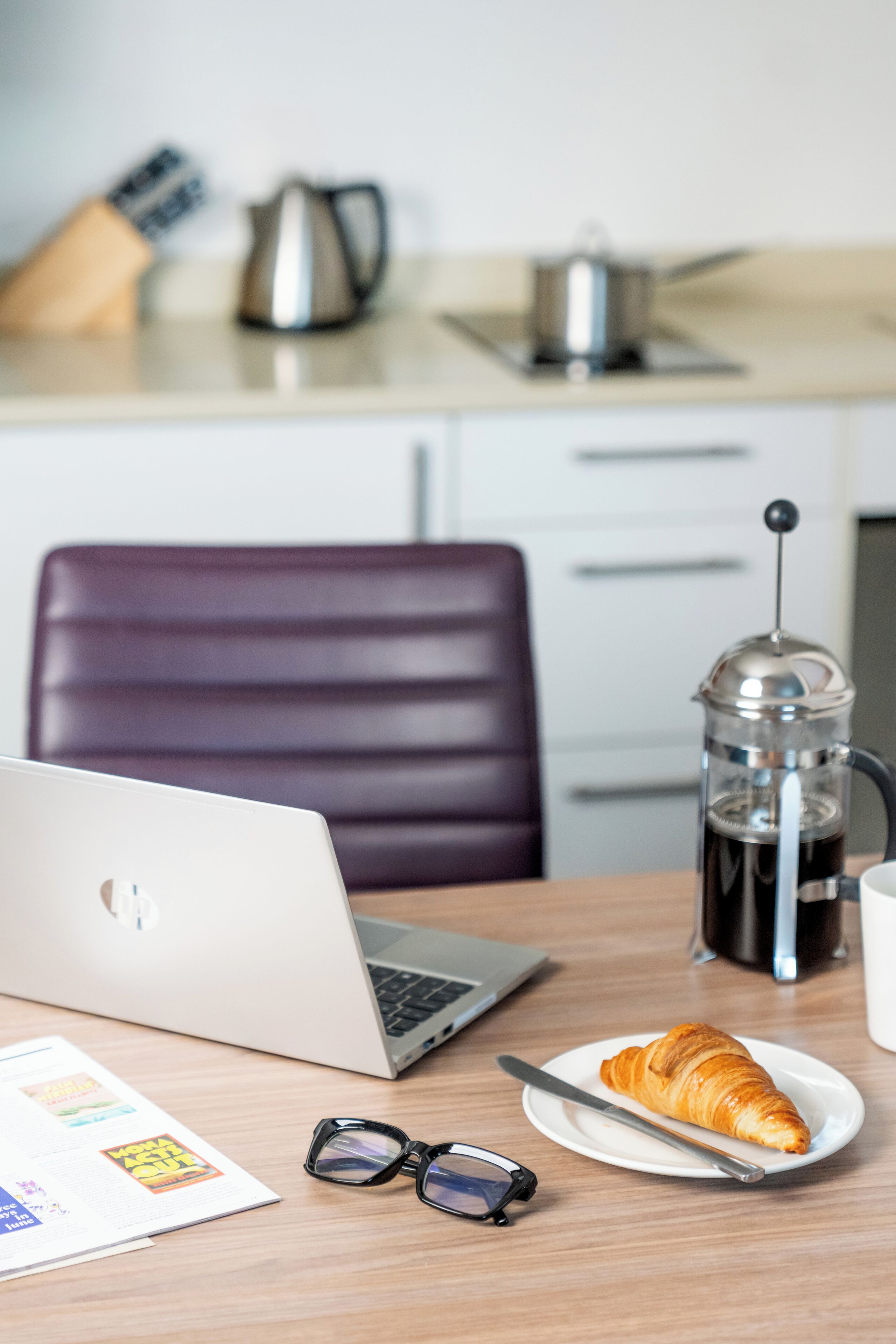 Ein Laptop, eine Kaffeemaschine, ein Teller mit einem Croissant und eine Brille auf einem Tisch.