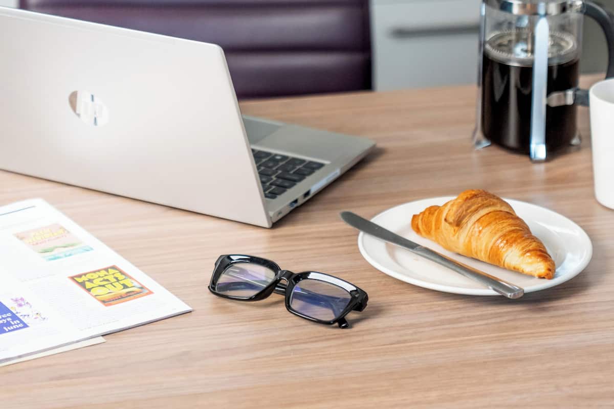 Kannettava tietokone, kahvinkeitin, lautanen, jossa on croissant, ja silmälasit pöydällä.