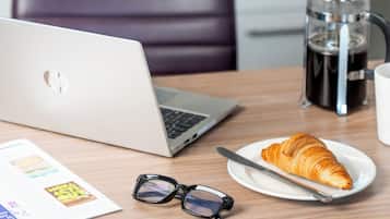 Una laptop, una cafetera, un plato con un croissant y un par de lentes sobre una mesa.