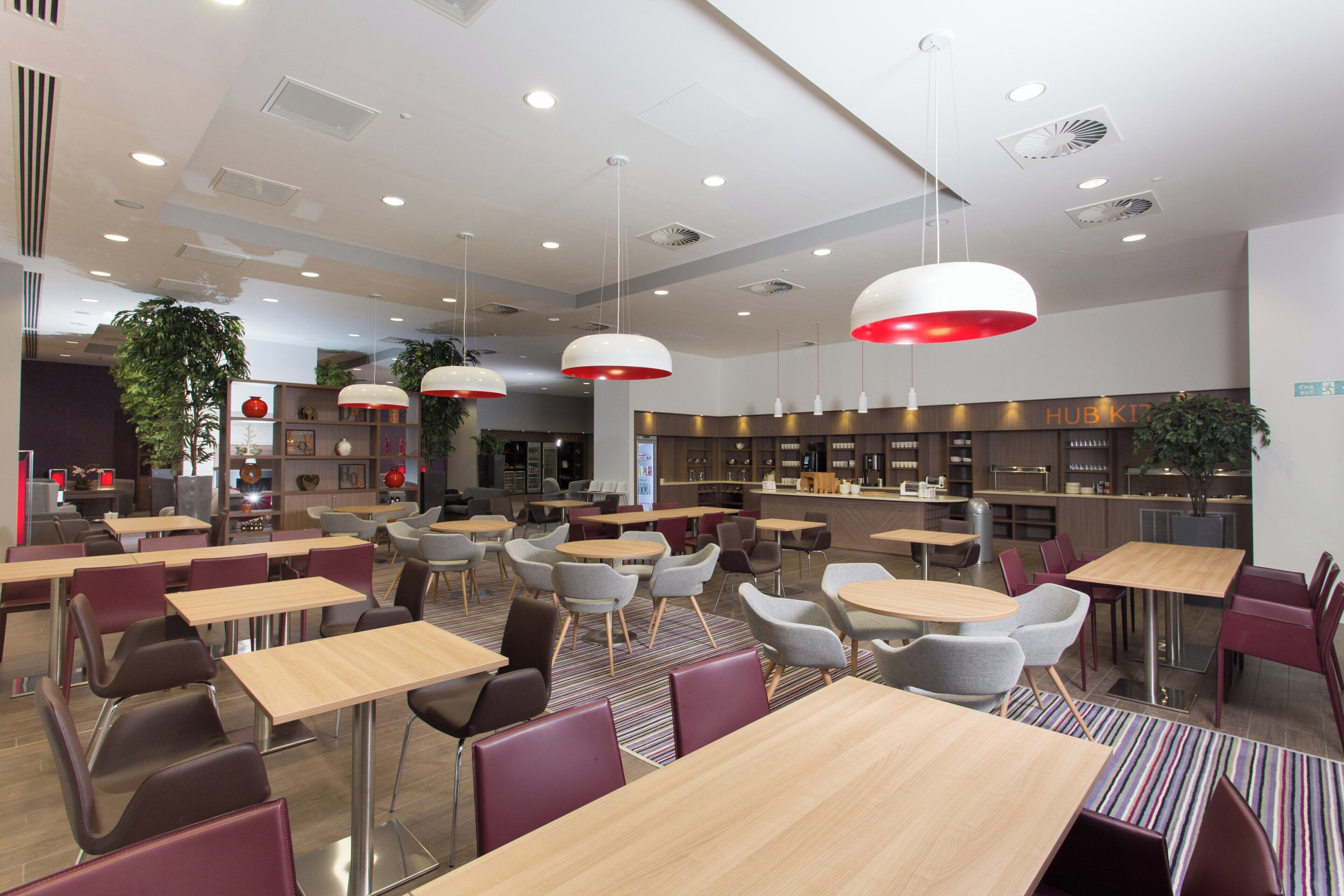 A modern dining area with multiple tables and chairs, overhead lighting, and a kitchen area in the background.