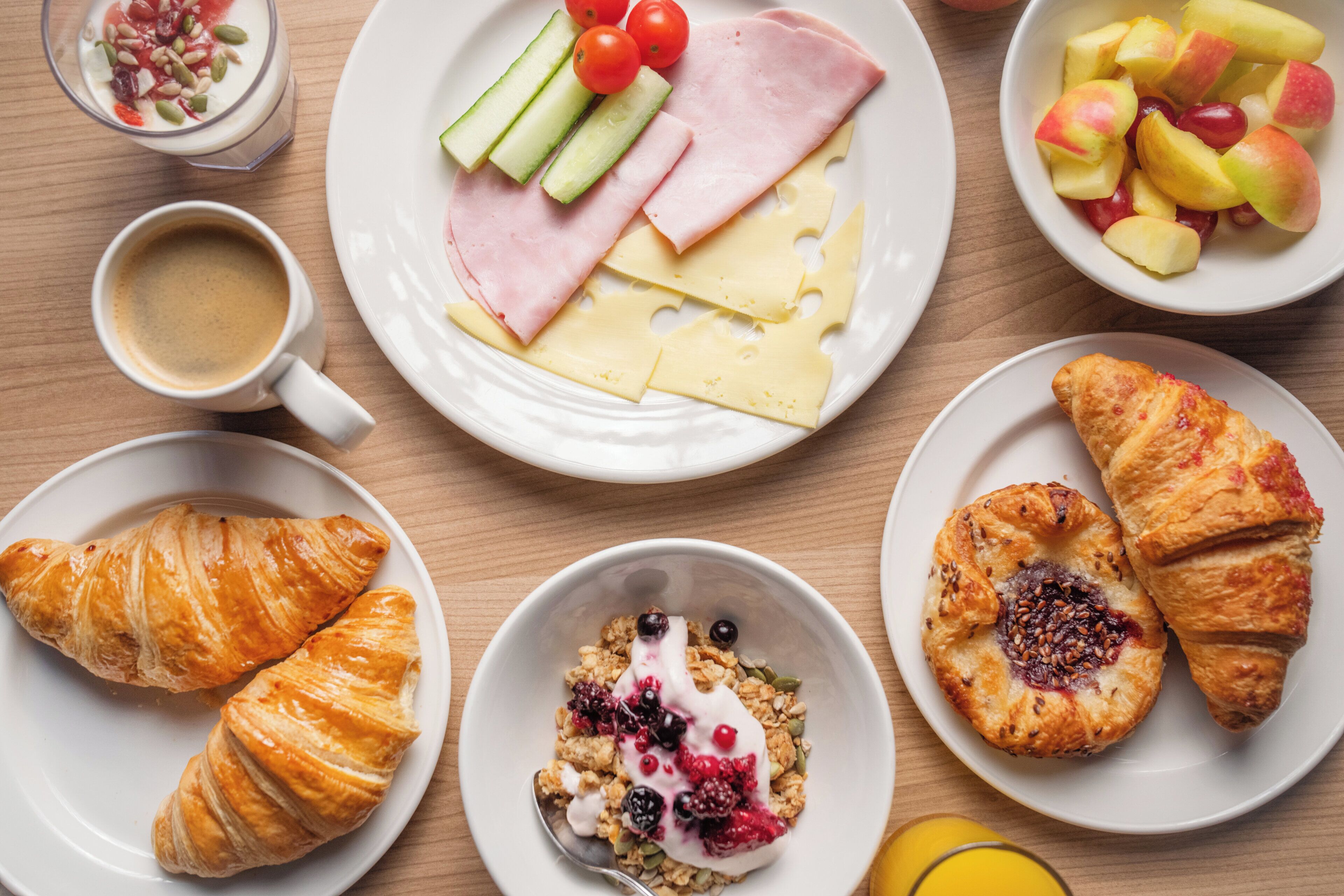 A breakfast setting with croissants, cheese, ham, yogurt with berries, and fruit on plates.