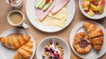 Ein Frühstückstisch mit Croissants, Käse, Schinken, Joghurt mit Beeren und Obst auf Tellern.