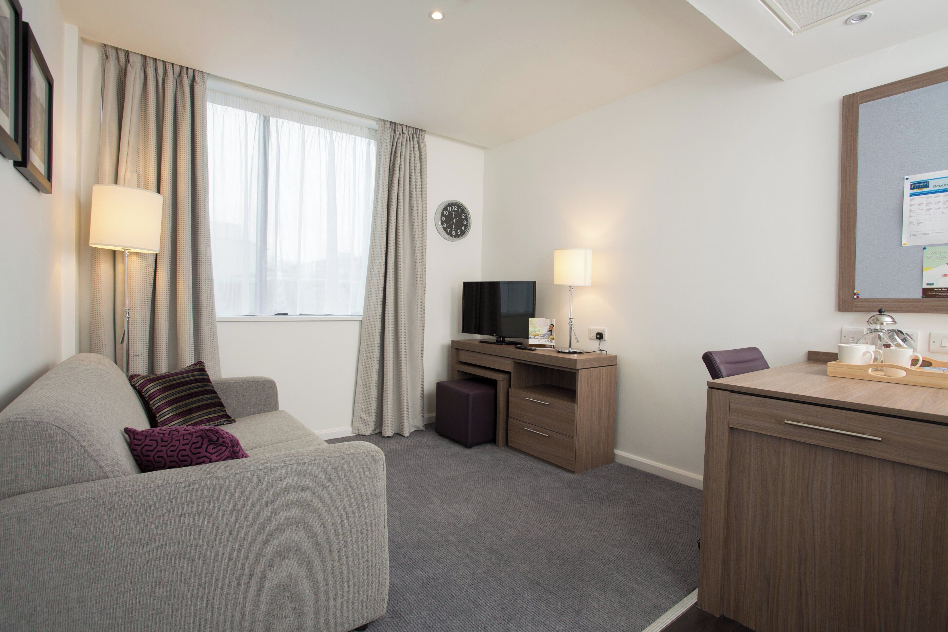 Habitación de hotel con sofá, escritorio, computadora, televisor, reloj y espejo.