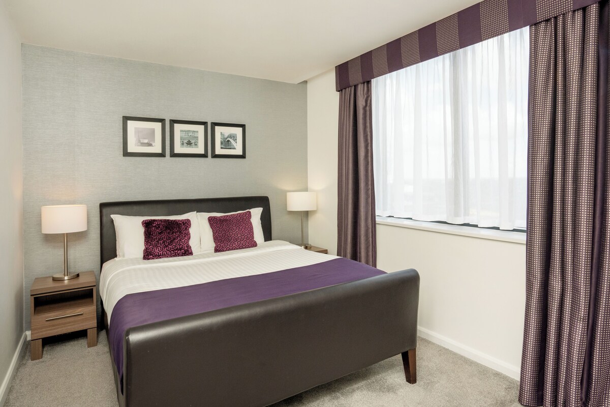 A neatly arranged hotel room with a large bed, a bedside table with a lamp, and a window with curtains.