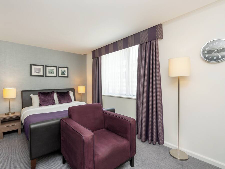 A hotel room with a bed, a chair, a bedside table with a lamp, a clock on the wall, and curtains.