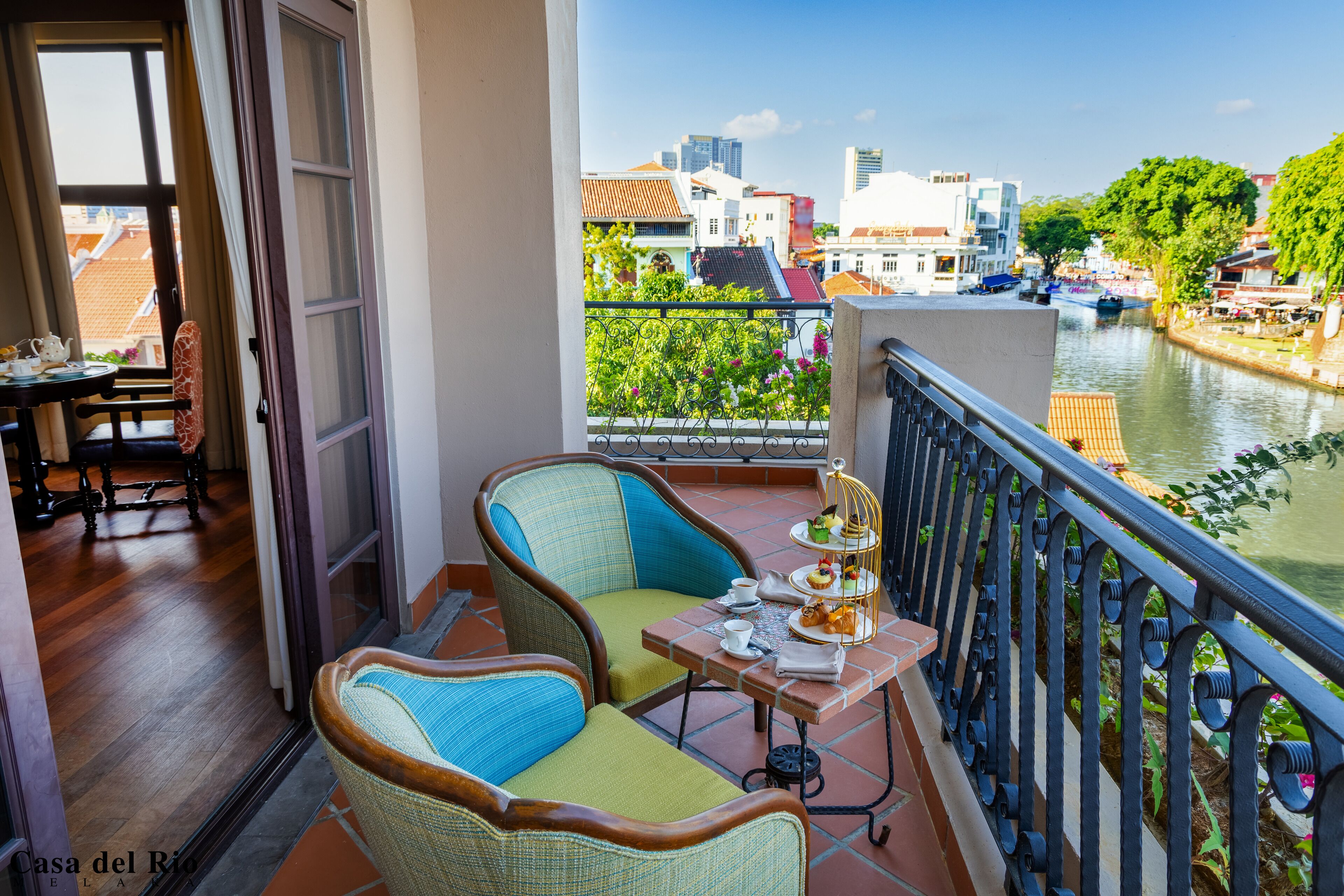 A balcony with wicker chairs, a small table with a tray of pastries, and a view of a river with boats.