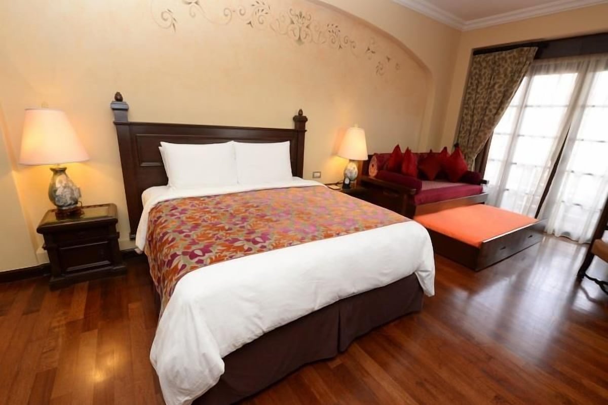 Una habitación de hotel con una cama grande, dos mesitas de noche con lámparas, un sofá rojo y una ventana con cortinas.
