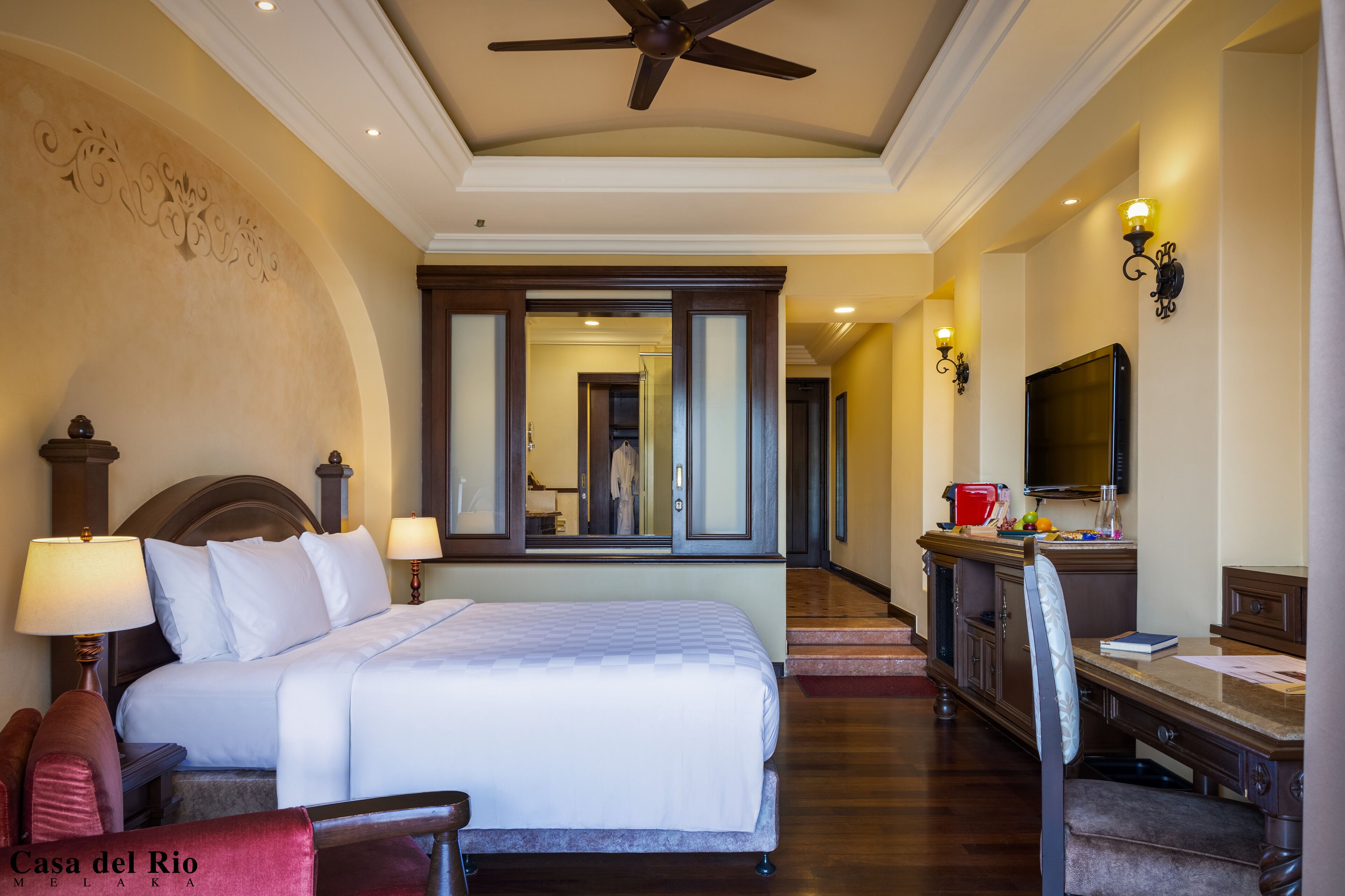 A hotel room with a large bed, a desk with a chair, a TV, and a ceiling fan.